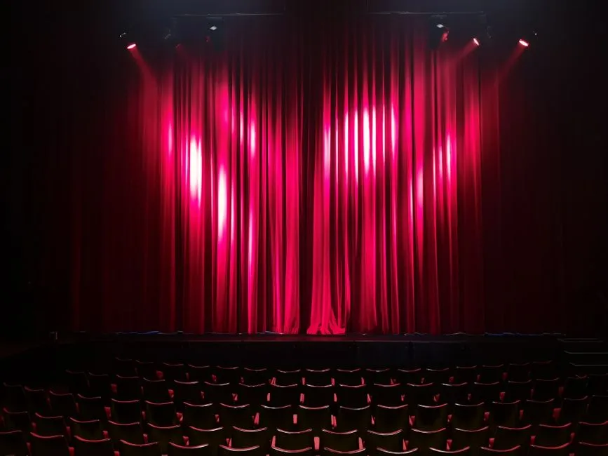 A dramatically lit red stage curtain with spotlights above rows of empty theatre seats in the foreground