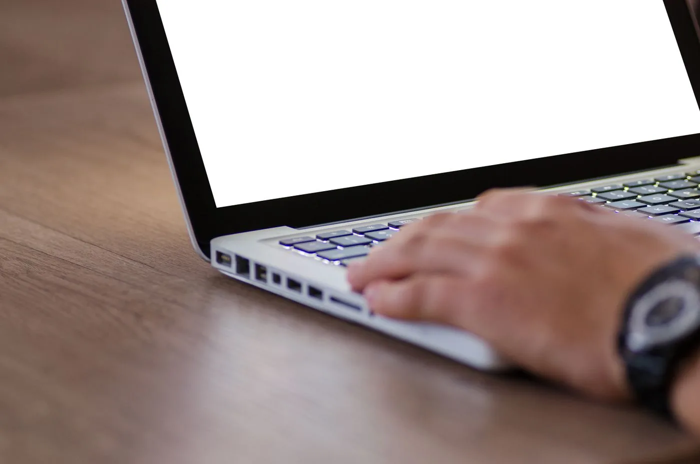 Close-up of a hand typing on a laptop keyboard with a blank white screen, on a wooden desk