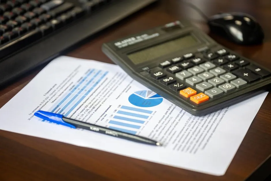A calculator resting on a printed financial report with a bar chart and pie chart, next to a keyboard and computer mouse on a wooden desk