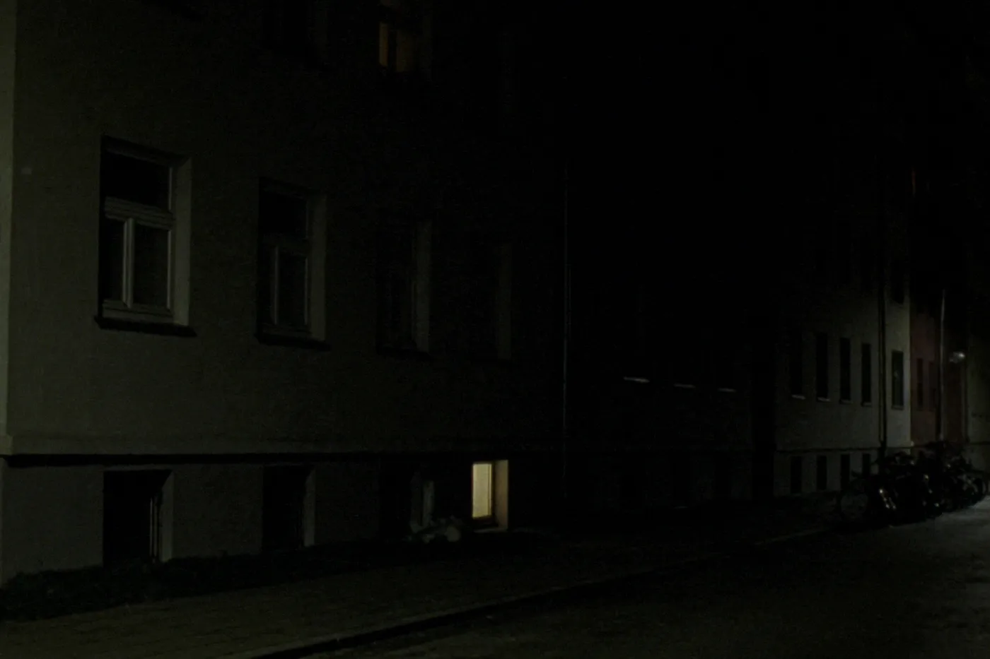 A single softly lit basement-level window glowing warm yellow along an otherwise dark row of apartment buildings on an empty residential street at night, a few bicycles parked far down the street, no people visible