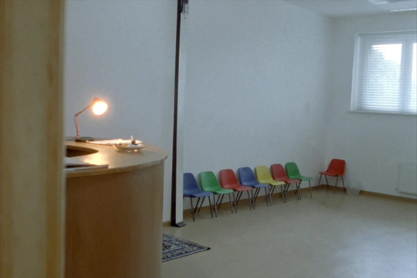 A calm, empty pediatric practice waiting area with a row of small colorful chairs against a plain wall and a warm reception desk lamp glowing nearby
