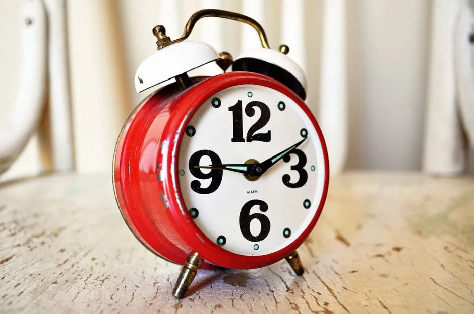 A red vintage double-bell alarm clock with its hands pointing to two o'clock, sitting on a wooden surface, no people visible