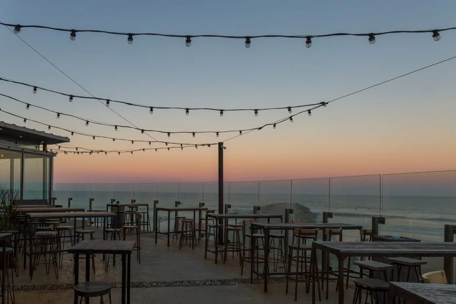 An empty rooftop terrace at dusk with string lights strung overhead, empty bar-height tables and stools, and a glass railing overlooking the sea and sky at sunset
