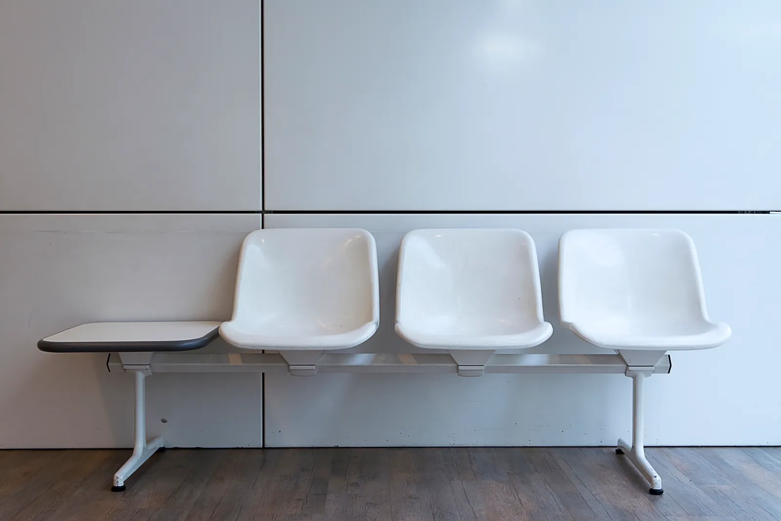 Three empty white plastic bench chairs against a plain white paneled wall, no people visible