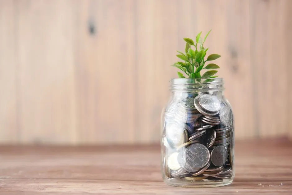 A glass jar filled with coins with a small green plant growing out of the top, sitting on a wooden table