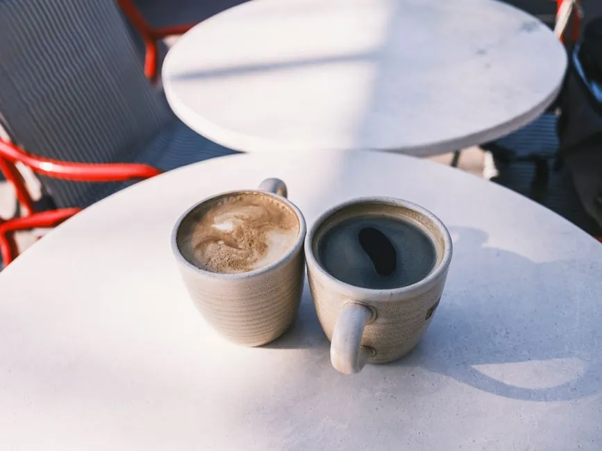 Two ceramic coffee cups side by side on a round outdoor cafe table in warm sunlight, one topped with milk foam, empty chairs visible at a neighboring table