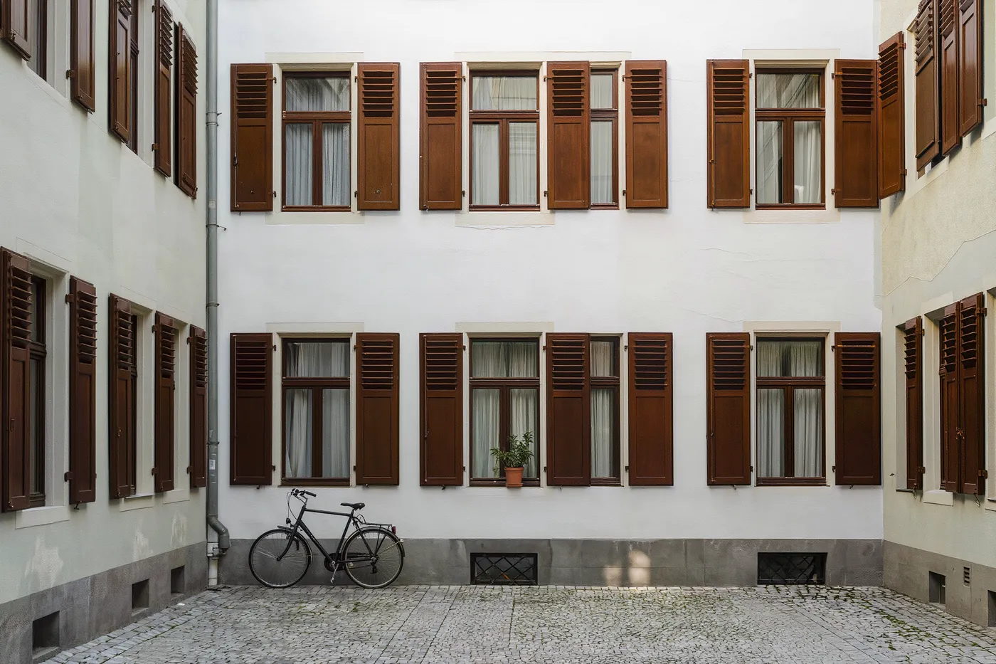 A quiet Berlin Altbau courtyard at midday, closed wooden window shutters, a bicycle leaning against the wall, a potted plant on a windowsill, empty cobblestones