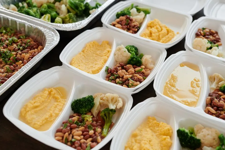 Rows of portioned lunch trays with vegetables and grains being plated for bulk school catering delivery