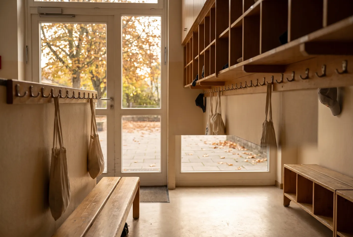 A bright, empty primary school entrance hallway in autumn, rows of wooden coat hooks and cubbies along the wall, autumn leaves visible through a glass door, no people or readable text