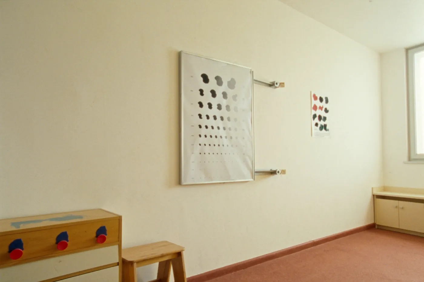 A bright, empty pediatric health office exam room, a height chart and an abstract vision test poster on a pale wall, a small wooden step stool and a low cabinet, warm daylight through a window, no people or readable text