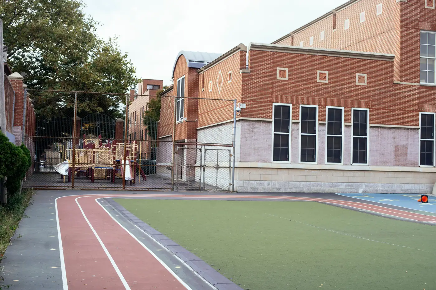 A contemporary red brick school building with a fenced running track and grassy sports ground, playground equipment visible behind a chain-link fence, no people or readable text or logos
