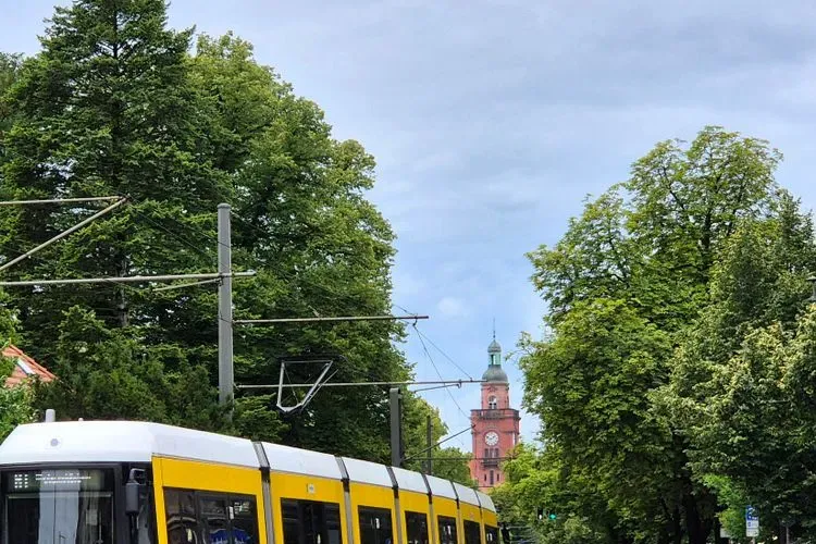 A yellow BVG tram running on overhead wires past trees, with a red brick clock tower visible in the background on a Berlin street
