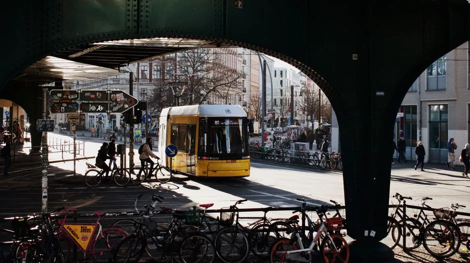A yellow Berlin tram passing beneath a railway bridge on a sunlit street, with rows of parked bicycles in the foreground and two cyclists riding alongside the tram
