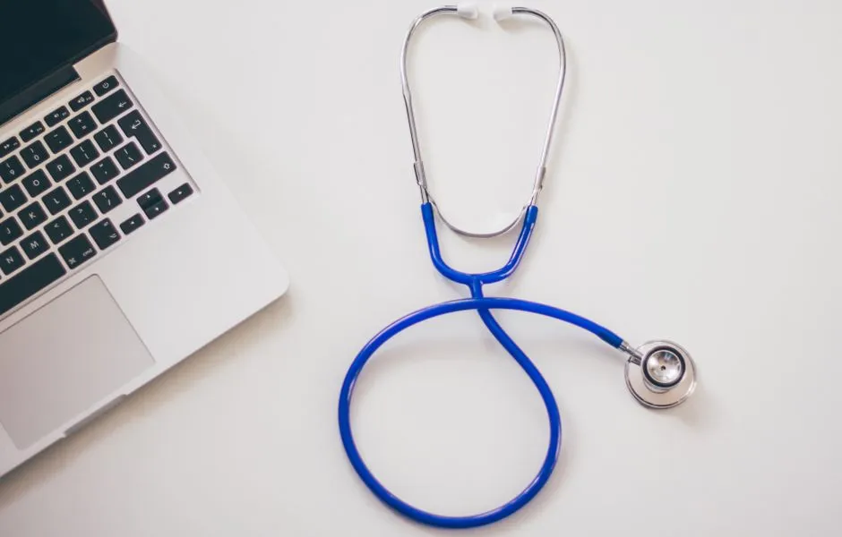 A blue stethoscope resting beside a laptop keyboard on a plain white desk, no people or readable text visible