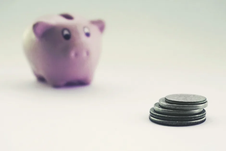 A small stack of coins beside a blurred pink piggy bank on a plain white surface