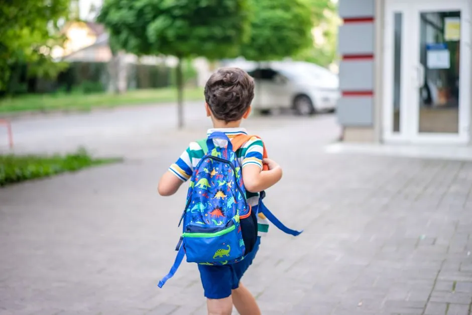 A child with a colorful backpack walking alone toward a school building on a paved courtyard