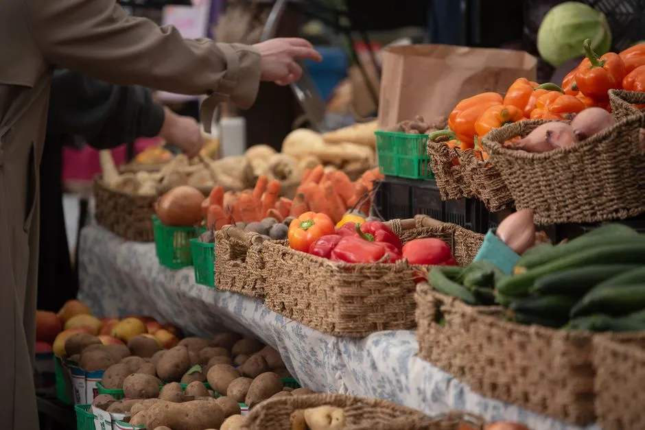 A market vendor's arm reaching over woven baskets piled with fresh peppers, carrots, potatoes, and cucumbers on an outdoor stall table, no face visible