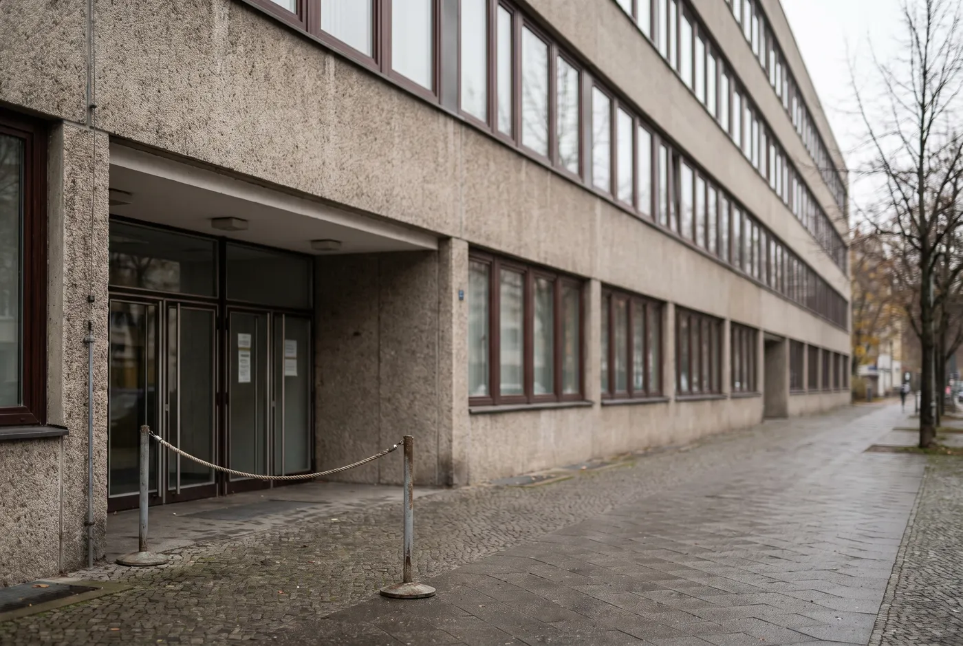 Exterior entrance of a Berlin district administration building with a plain postwar facade, an empty pedestrian queue barrier near the doorway, and no readable signage visible