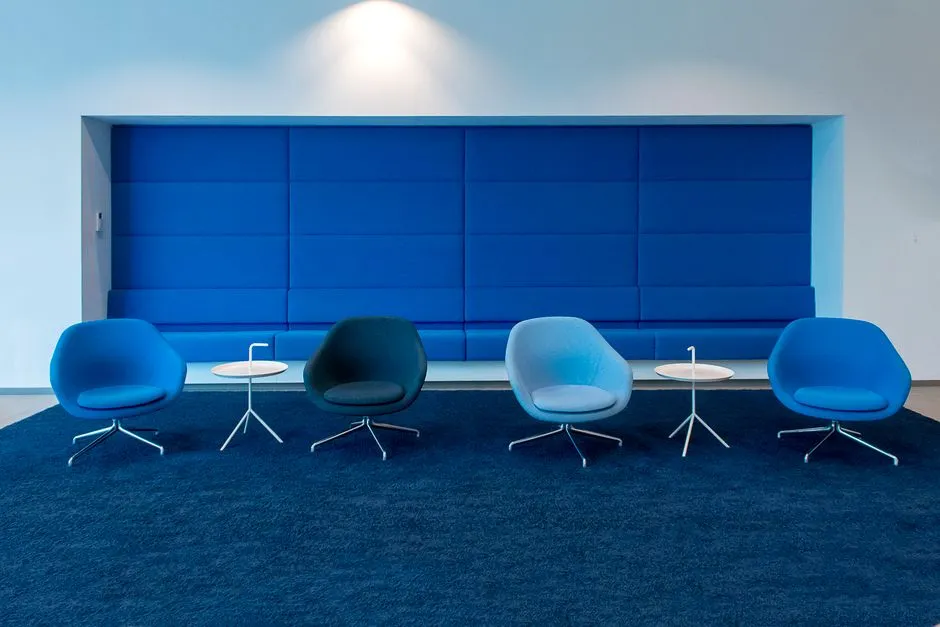 A modern office waiting area with several blue lounge chairs arranged in a row facing a padded blue wall panel, small round side tables between them, on a dark blue carpet, no people or readable text visible