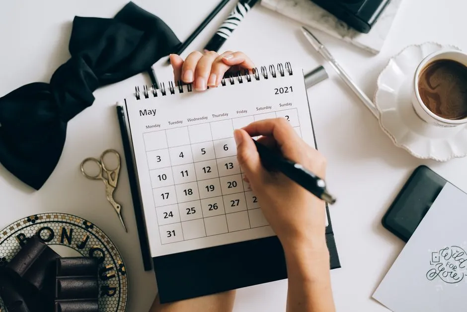 Una mano marcando una fecha en un calendario de escritorio de papel, con una taza de café y un teléfono cerca