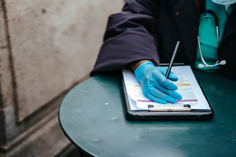 A gloved medical worker's hand writing on a clipboard of paperwork, with a stethoscope draped over the shoulder
