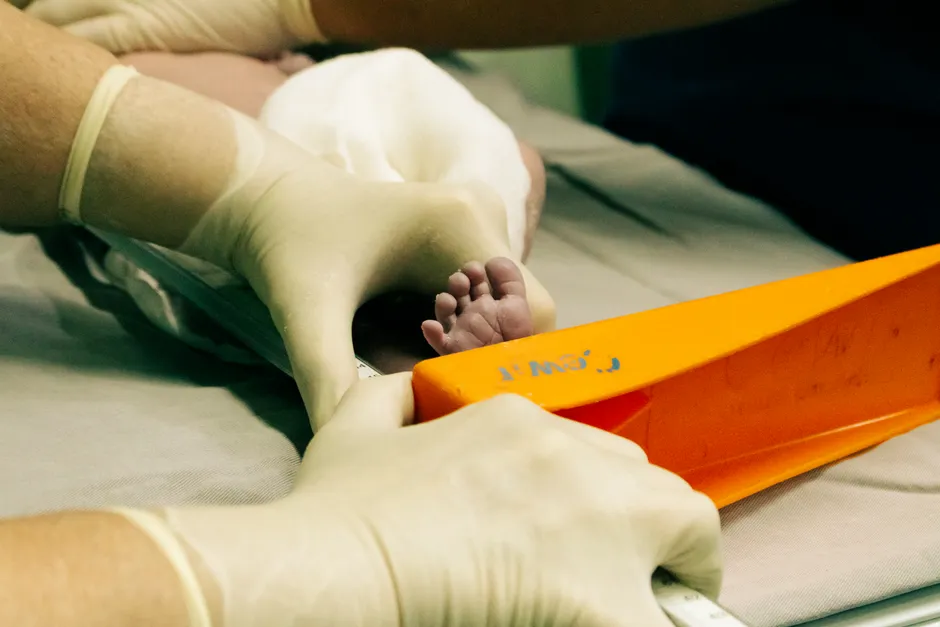 Der Fuß eines Neugeborenen wird sanft von behandschuhten Händen in einer Klinikumgebung gehalten, kein Gesicht sichtbar