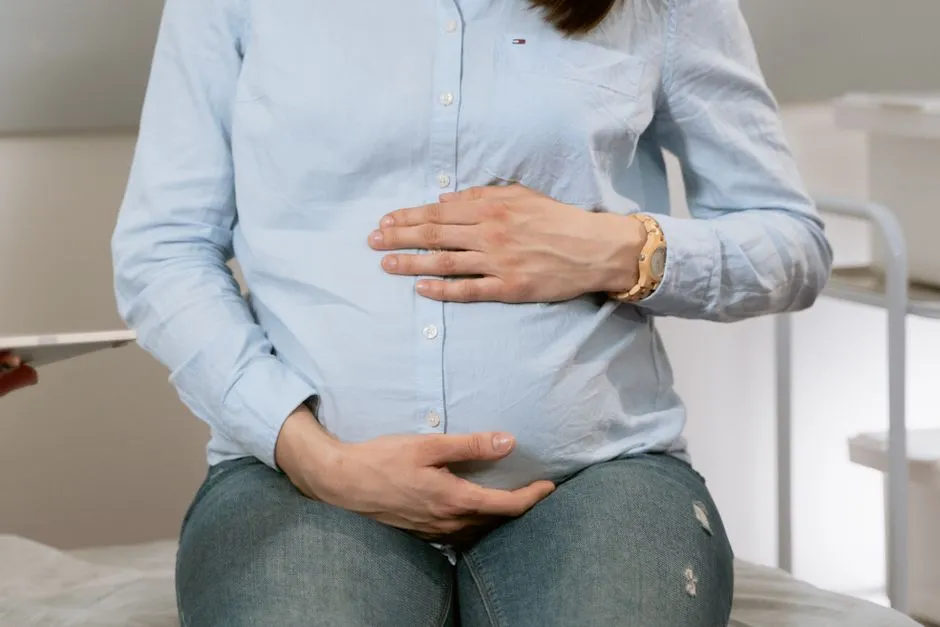 A pregnant woman in a light blue shirt resting her hands on her belly while seated