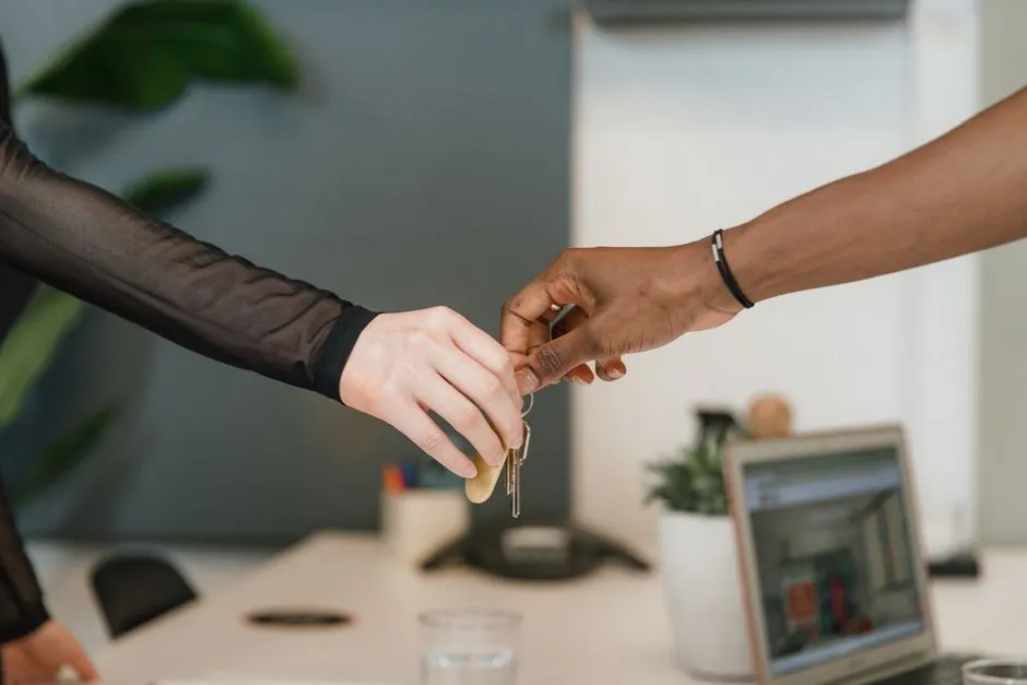 Two hands exchanging a set of car keys across an office desk, with a laptop and a small plant in the background