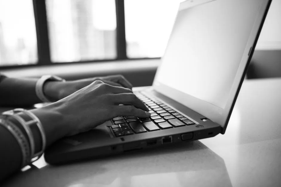 Una foto en primer plano y en blanco y negro de dos manos escribiendo en el teclado de un portátil junto a una ventana