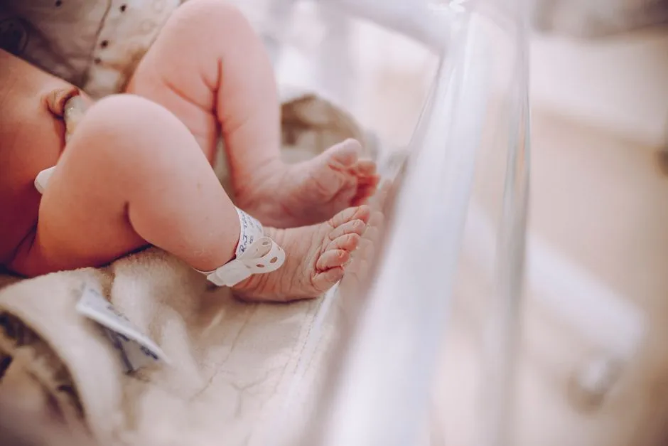A newborn's feet resting on a blanket in a hospital crib, with an identification band visible on the ankle