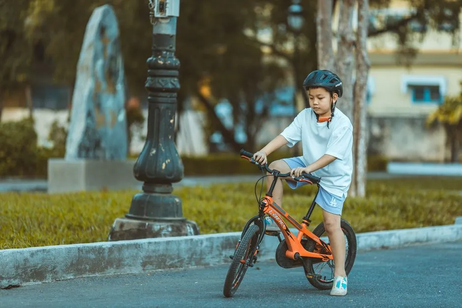 Ein Kind mit Fahrradhelm fährt auf einem orangefarbenen Fahrrad auf einem asphaltierten Weg in einem Stadtpark