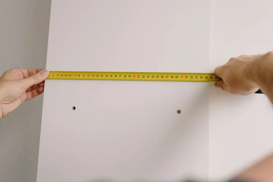 Two hands holding a yellow tape measure between two existing drill holes on a white wall, checking the spacing before hanging something new