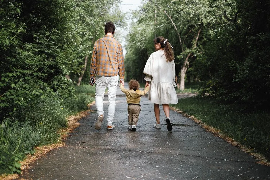 A parent, another adult, and a young child walking away down a tree-lined path, seen from behind, no faces visible