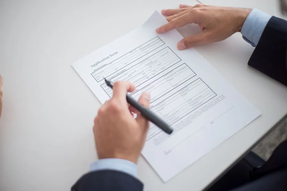 Two hands over a printed application form on a desk, one pointing with a pen, no faces visible