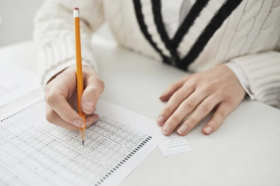 A close-up of two hands at a table, one holding a pencil marking bubbles on a multiple-choice answer sheet, the other resting flat on the desk, no face visible