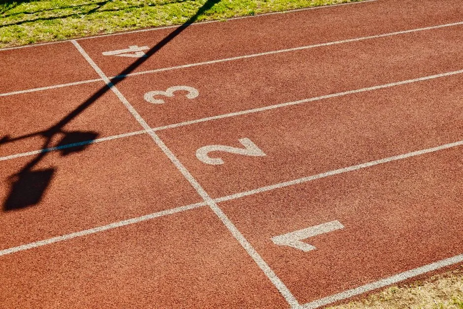 An empty outdoor running track with white lane-number markings one through four painted on a reddish-brown rubber surface, a faint shadow of a lamp post crossing the lanes, green grass visible at the edge, no people in view