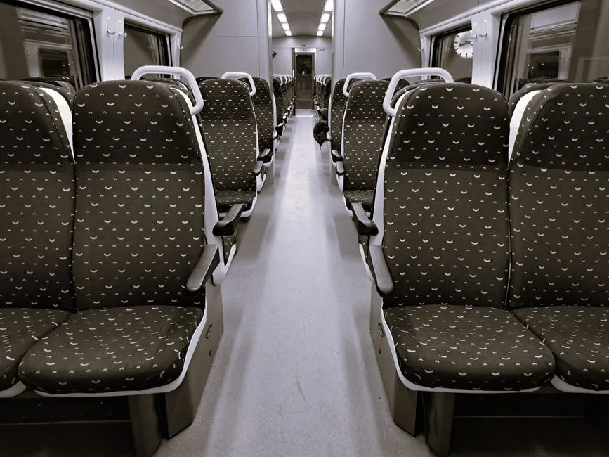 Rows of empty patterned train seats facing each other down the aisle of a regional train carriage, seen from the center of the car looking toward the far end, no people, no readable text or logos visible