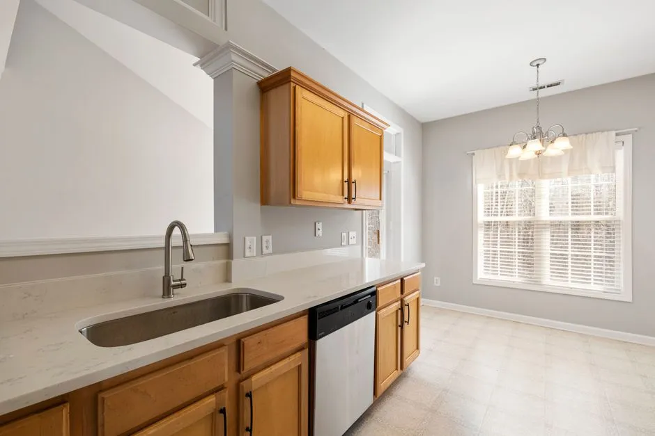 Empty finished kitchen with wooden cabinets, marble-look countertop, stainless steel dishwasher, and a sink, dining nook with window visible in the background