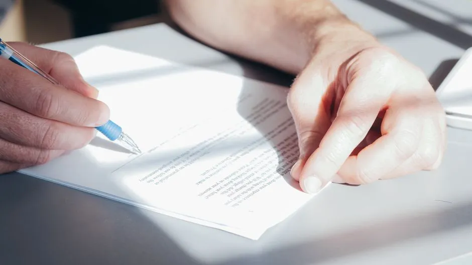 Two hands holding a pen over a partially signed legal document on a desk