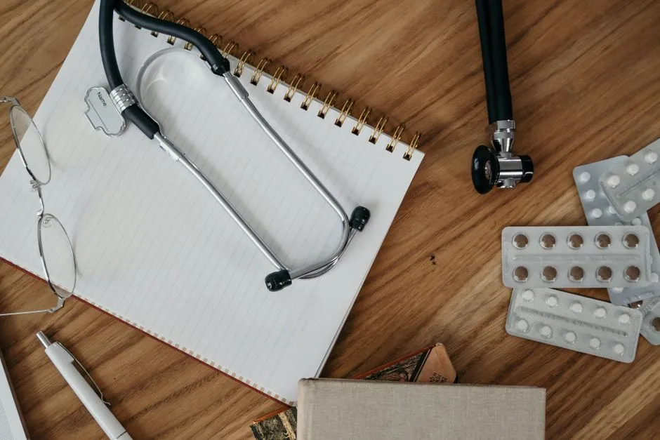 A stethoscope, glasses, and blister packs of pills laid out on a spiral notepad on a wooden desk