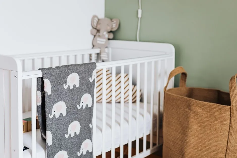 A white wooden crib with a gray elephant-print blanket draped over the rail, a small plush elephant toy, and a woven storage basket beside a sage green wall