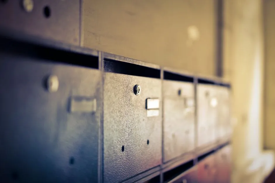 A row of weathered metal apartment mailboxes seen at a low angle, each with a blank nameplate and a narrow mail slot