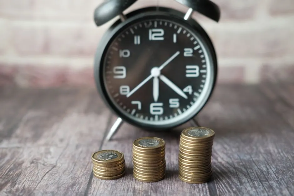 A black alarm clock behind three ascending stacks of gold coins on a wooden table