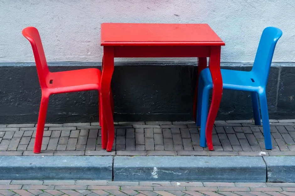 Two empty plastic chairs, one red and one blue, positioned facing a small red table against a plain wall on a brick sidewalk, no people in view