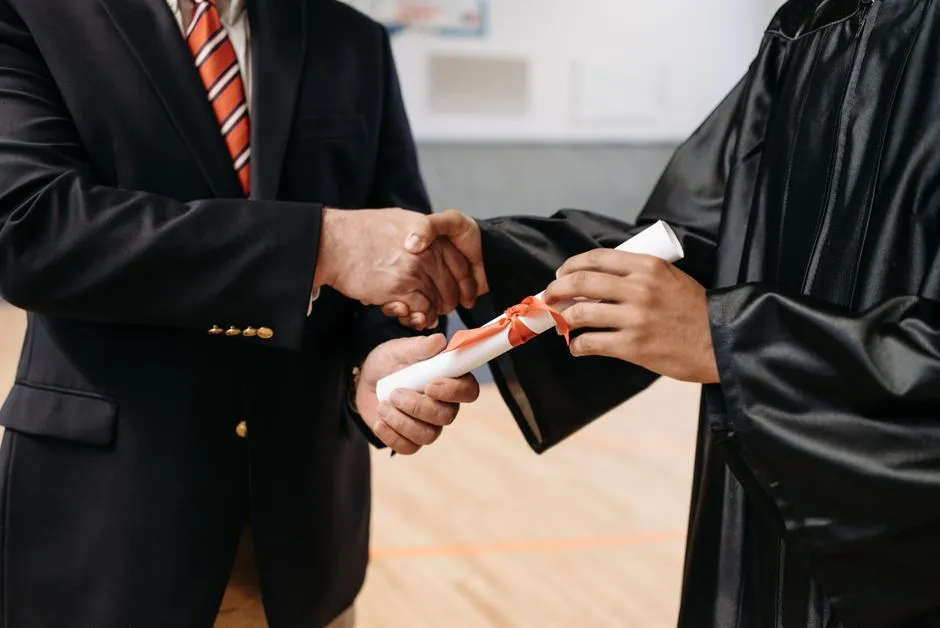Two people in formal attire shaking hands while one hands over a ribbon-tied rolled certificate, close-up on the handshake and document, no faces visible