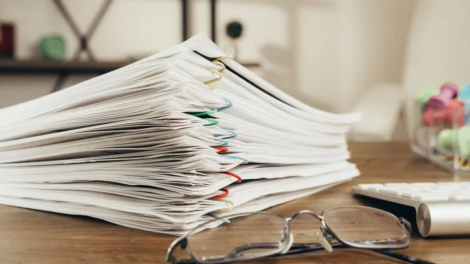 A thick stack of paper-clipped documents on a wooden desk, next to a pair of reading glasses and the edge of a keyboard, no text legible