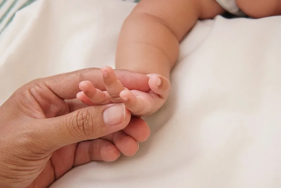 A parent's hand gently holding a newborn baby's small hand and fingers on a soft white blanket