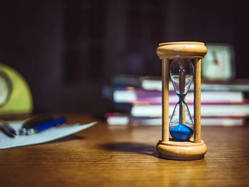 A wooden hourglass with blue sand on a desk, with a blurred stack of books and a notepad with a pen in the background