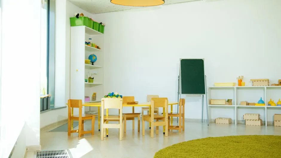 An empty, bright kindergarten-style classroom with small wooden chairs and a low table, a green chalkboard, open shelving with wooden toys and a globe, no people or readable text