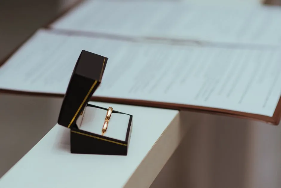 A wedding ring in an open jewelry box on a desk, blurred official documents in the background, no readable text or faces visible